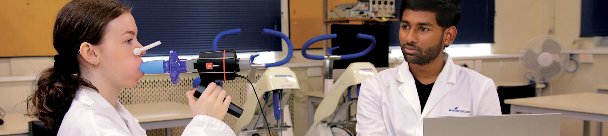 A photo of two students in the lab; one student breathes into a spirometer while the other student looks on.